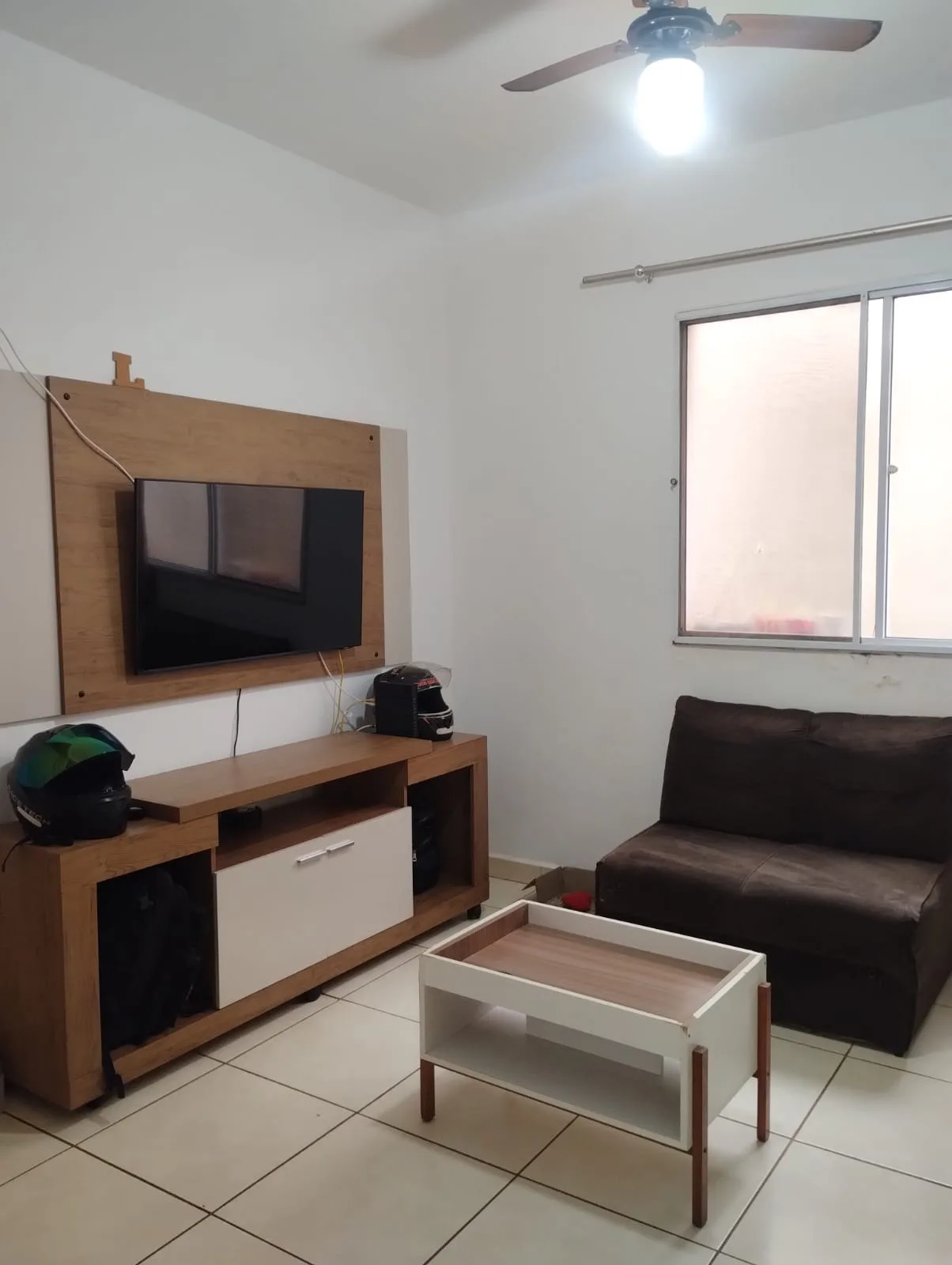 Sala de estar com painel de TV, mesa, sofá, janela e ventilador de teto fotografados.