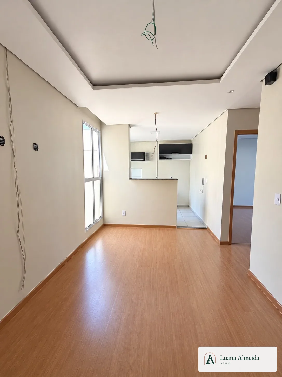 Sala vazia do apartamento com piso amadeirado, janela lateral e balcão da cozinha ao fundo.